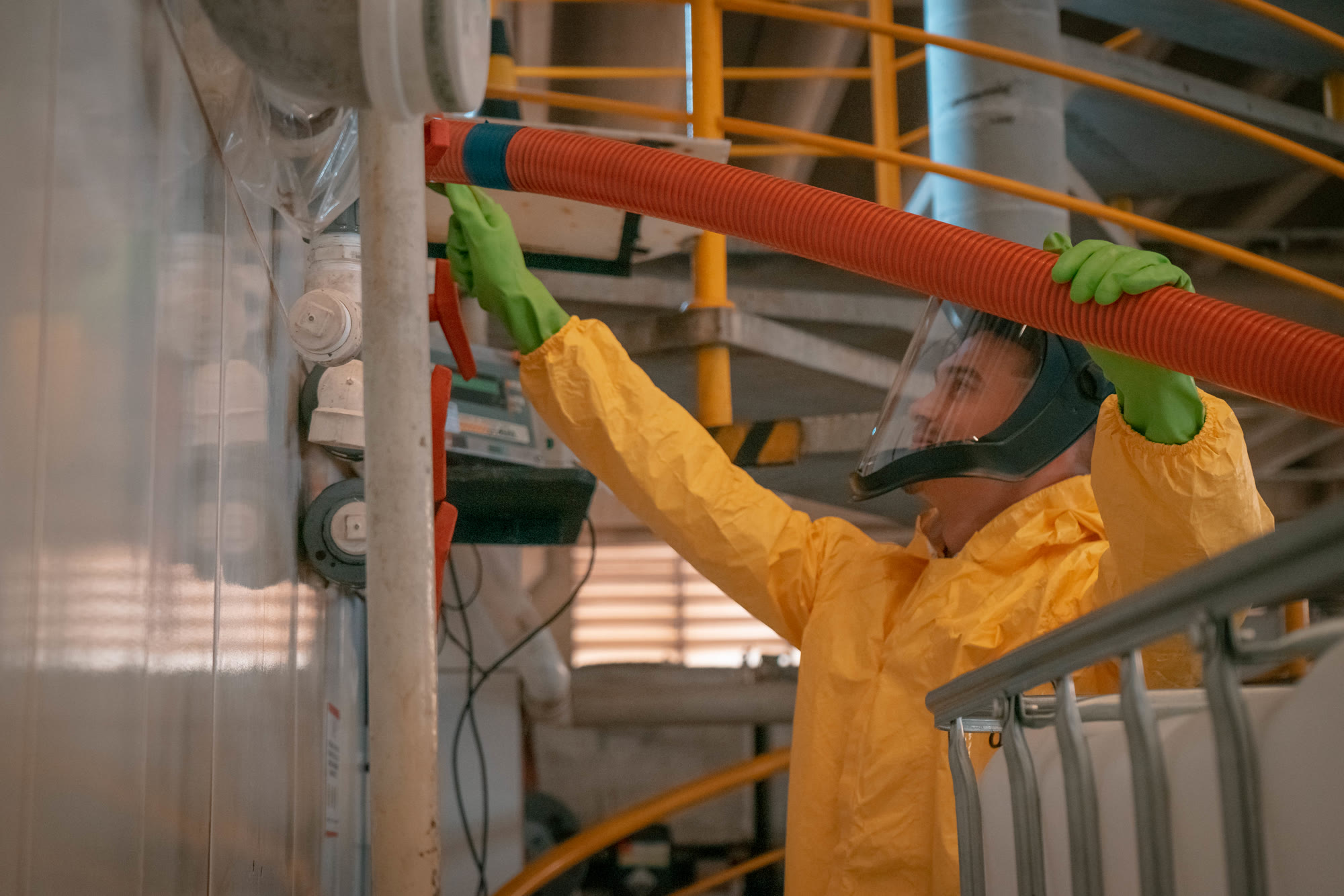 Colaborador da Hanaoka com traje de proteção completo (macacão amarelo, luvas e protetor facial) realizando manutenção em equipamento na planta industrial
